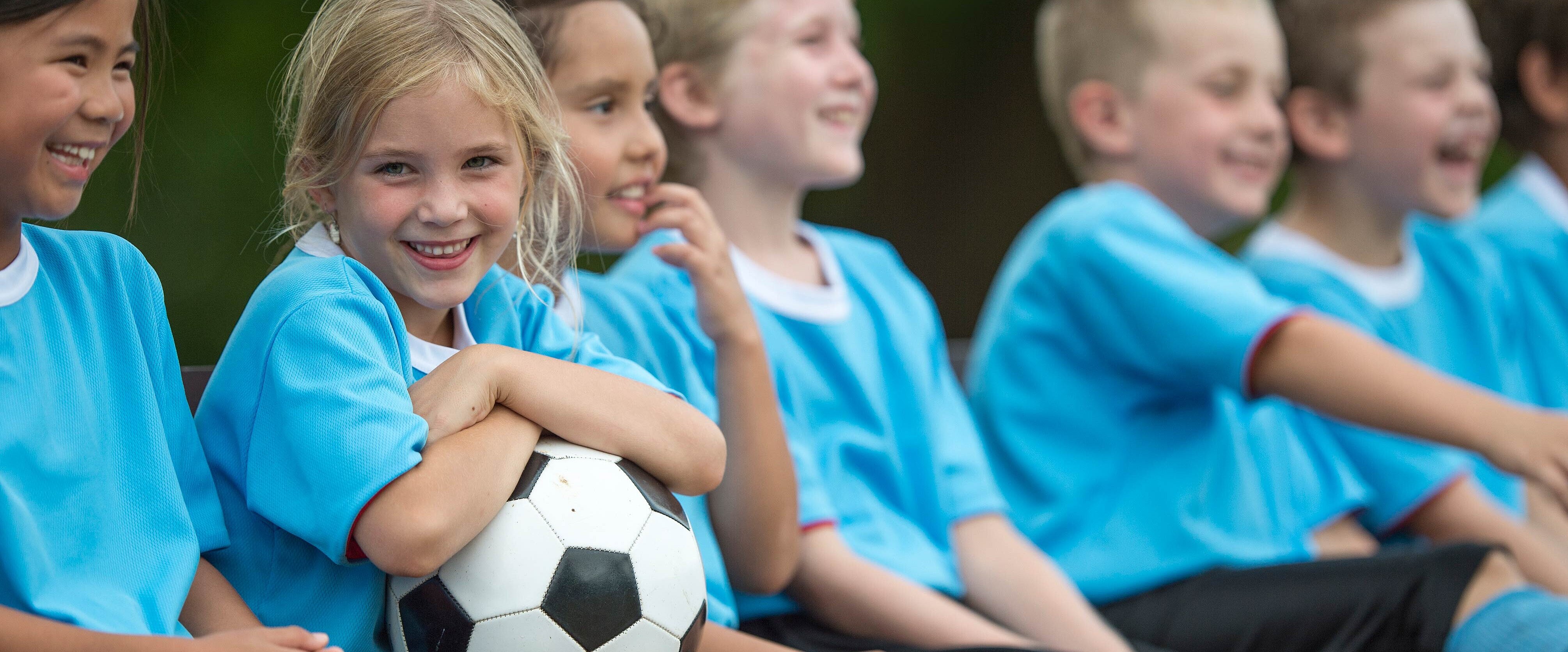 Möglichst vereint Sieben Kinder sitzend in hellblauen Fußballtrikots, ein Mädchen hält den Fußball
