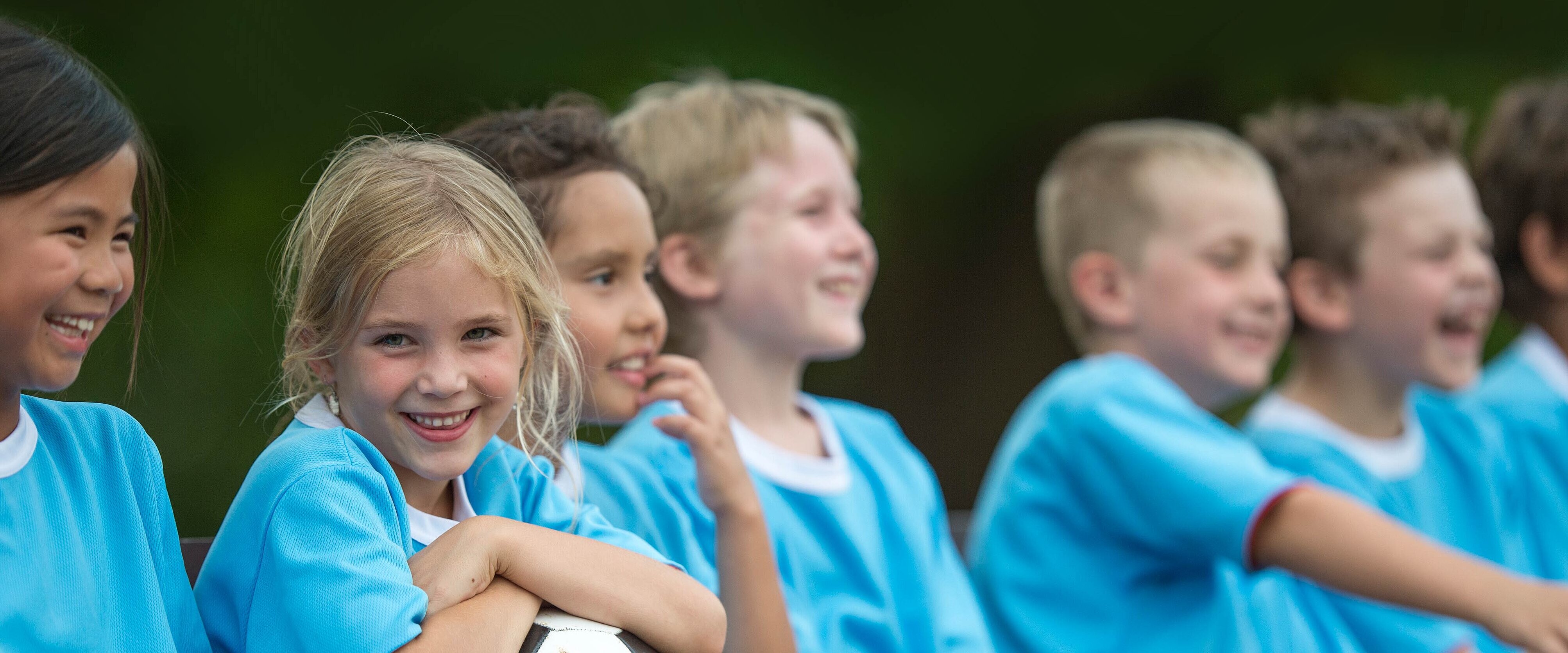 Kindergruppe, die Fußballtrikots trägt und in einer Reihe neeneinander sitzt.