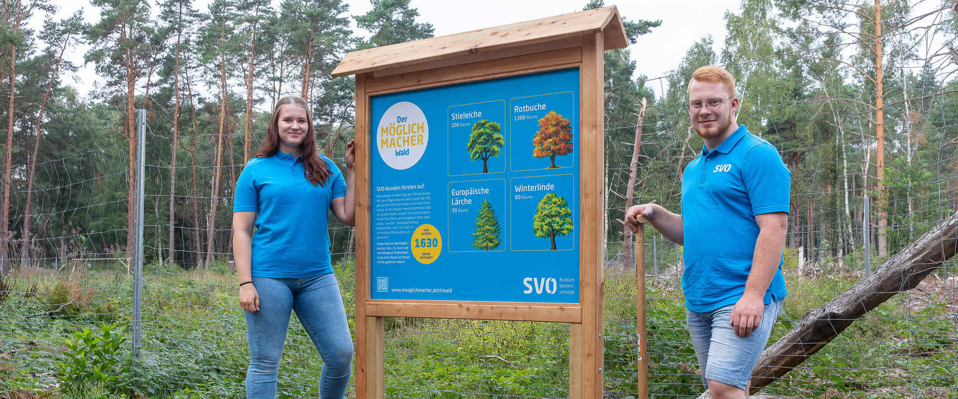 Zwei SVO-Mitarbeitende stehen neben einer Informationstafel über Baumarten im Wald.